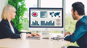 Zwei Personen sitzen vor einem Bildschirm und besprechen Daten, Grafiken, Statistiken 