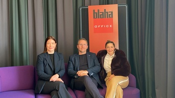 Drei Personen sitzen auf einem violetten Sofa vor einem Vorhang; im Hintergrund steht ein Schild mit der Aufschrift „blaha OFFICE“, am Boden liegt ein gemusterter Teppich.