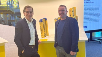 Zwei Personen stehen an einem gelben Messestand mit der Aufschrift „Formwork beams | Schalungsträger“, im Hintergrund eine Messehalle mit Gerüsten und Monitor.