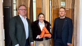 Drei Personen stehen in einem Innenraum vor einer Präsentationswand mit verschiedenen Holzoberflächen; eine Person hält eine dreieckige, orangefarbene Auszeichnung in den Händen.
