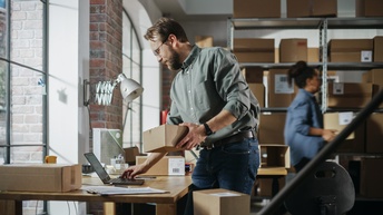 Personen arbeiten im Lagerraum am Laptop, bereiten Pakete für die Auslieferung vor und verpacken sie.