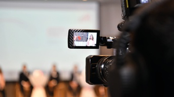 Blick von hinten durch eine Kamera auf eine Vortragende bei einem Webinar
