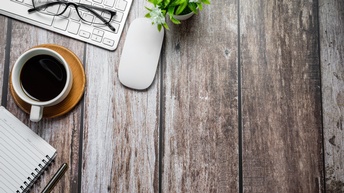 Perspektive von oben auf einen Holzuntergrund mit Kaffee, Tastatur, Maus, Brille sowie Notizbuch und Stift