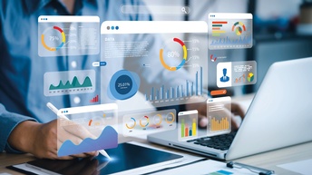 Analyst arbeitend an einem Computer und Tablet. Er analysiert Geschäftsdaten dargestellt mit bunten Statistiken und Matriken.<br />
