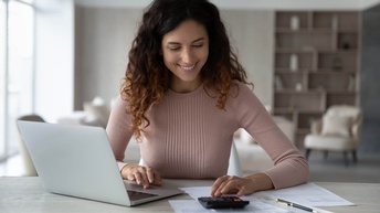 Frontalaufnahme einer Person, die hinter einem Tisch sitzt. Rechts vor der Person ist ein aufgeklappter Laptop. Links vor ihr ist ein Taschenrechner, auf den sie blickt. Unter und um den Taschenrechner sind bedruckte Zettel