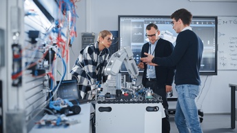 Drei Personen stehen in einem Raum mit Kabeln auf Schienensystemen. Sie unterhalten sich konzentriert über einen Roboterarm, der vor ihnen auf einem Tisch steht. Im Hintergrund ist ein großer Wandbildschirm mit Diagrammen.