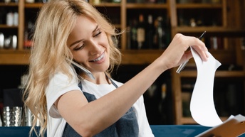 Person mit langen blonden Haaren und Schürze telefoniert mit einem Smartphone in der Schulter eingeklemmt und steht vor einer Schank und blickt auf Dokumente auf einem Klemmbrett