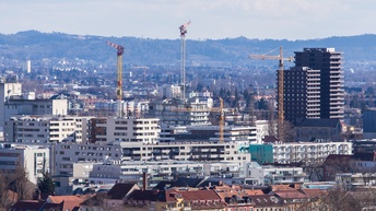Panoramabild der Stadt Graz mit Häusern, Neubauten, Baustellenkränen und im Hintergrund befindet sich ein angrenzendes Waldgebiet