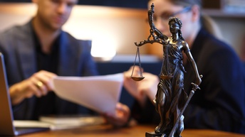 Im Bildvordergrund  steht eine bronzene Statue, die eine Waage in der Hand hält und verbundene Augen hat. Im Hintergrund sitzen zwei Personen an einem Tisch. Die linke Person hält einen Zettel. Beide blicken darauf