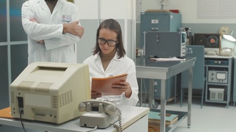 Eine Person in weißer Arbeitskleidung in einem Labor sitzt an einem Tisch mit einem Computer und einem Telefon und blickt dabei auf ihre in den Händen gehaltenen Unterlagen, eine zweite Person in weißer Arbeitskleidung steht mit verschränkten Armen hinter ihr