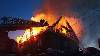 Ein brennender Dachstuhl eines Wohnhauses mit Flammen, Funken und aufsteigendem Rauch vor dem Abendhimmel. Links neben dem Haus stehen auf einer ausfahrbaren Leiter eines Fahrzeugs zwei Personen