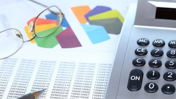 Nahaufnahme von Zahlenreihen - dahinter eine Brille und ein Taschenrechner