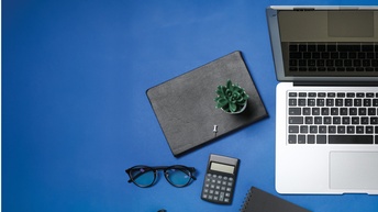 Laptop, Notizbücher, Taschenrechner, Brille sowie Stift, Büroklammer liegen auf einem blauen Untergrund, Topshot