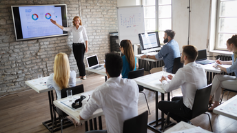 Rückenansicht von sechs Personen, die in einem Raum jeweils alleine an einem Tisch sitzen. Vor manchen Personen ist ein Laptop. Ihnen gegenüber steht eine Person an einem Bildschirm mit einer Grafik, auf die sie deutet