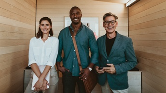 Drei lächelnde Personen stehen nebeneinander, im Hintergrund Holzwand