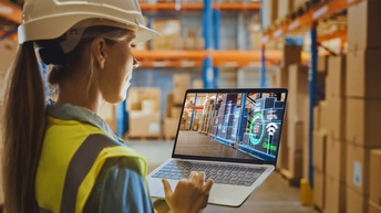 Person mit Helm und Schutzweste in Seitenansicht, die aufgeklapptes Notebook in Händen hält, auf Display des Notebooks Lagerhalle mit Paletten voller Kratons umrandet mit leuchtenden  Linien und Schriftzug 100% Efficiency