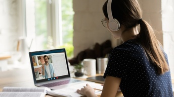 Rückenansicht einer Person mit Kopfhörern, die an einem Tisch sitzt. Direkt vor der Person ist ein Notizblock, daneben ist ein aufgeklapptes Buch. Vor dem Block steht ein Laptop, auf dessen Bildschirm eine Person in die Kamera spricht