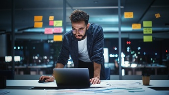Bärtige Person mit Brillen beugt sich über Tisch zu Laptop und betätigt Computermaus, im Hintergrund Glaswand mit bunten Post-Its