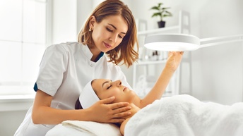 Eine Person ist über den Kopf einer  anderen Person gebeugt, die auf einem Massagebett liegt. Sie hält mit der linken Hand ihr Gesicht, mit der rechten Hand berührt sie eine Lampe, die über der liegenden Person schwebt