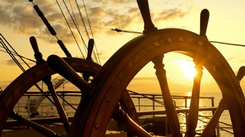 braunes Steuerrad eines Schiffes, im Hintergrund untergehende Sonne am Himmel, die durchs Steuerrad scheint