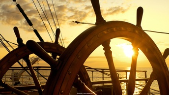 braunes Steuerrad eines Schiffes, im Hintergrund untergehende Sonne am Himmel, die durchs Steuerrad scheint