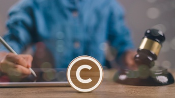 Ein in einem Kreis gefasstes Copyright-Zeichen steht als Icon auf einem Holztisch. Im Hintergrund ist eine Person, die etwas schreibt und einen Hammer in der Hand hält. Gerechtigkeit wird dargestellt.