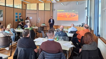 Raum mit mehreren Personen, die an Tischen sitzen, vorne steht ein Referent und hält eine Präsentation