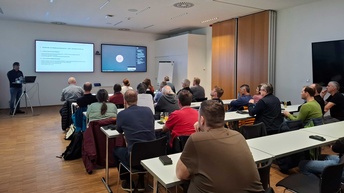 In einem Raum sitzen mehrere Menschen an Tischen und hören dem Referenten zu, der vorne steht