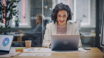 Person sitzt freudig bei einem Besprechungstisch und blickt auf einen aufgeklappten Laptop, hinter der Glaswand sitzt eine weitere Person bei einem Schreibtisch