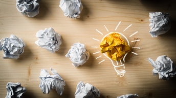 Auf einem Tisch liegen viele zerknäulte, weiße Zettel. Ein Zettel ist gelb. Um den Zettel sind die Umrisse einer Glühbirne illustriert