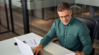 Geschäftsmann, der Dokumente bearbeitet, und dabei einen Laptop verwendet, sitzend an einem Schreibtisch <br />
