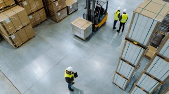 Topshot einer Lagerhalle, in der links und rechts viele, in Plastik gehüllte Kartons über- und nebeneinander gestapelt sind. Dazwischen ist ein Gabelstapler, mit dem eine Person fährt. Daneben stehen an verschiedenen Orten Personen mit Helmen 