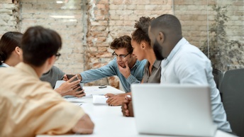 Sechs Personen sitzen sich an einem langen, hellen Tisch in einem Raum mit Glaswänden gegenüber. Eine Person links zeigt den rechts sitzenden Person etwas auf einem Laptop