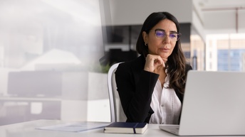 Eine Person mit langen und offenen Haaren sitzt mit einem ernsten Blick auf ihren Laptop in einem modernen Büro. Auf dem Tisch liegen ein dunkelblaues Notizbuch und mehrere Zettel.