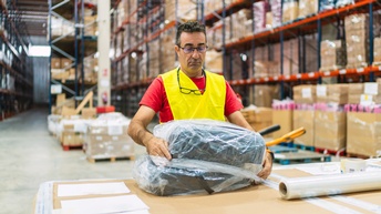 Eine Person steht an einem Tisch in einer Lagerhalle und verpackt Produkte in Plastikfolie.