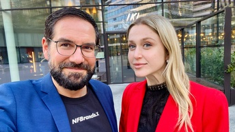 Porträt von zwei nebeneinander stehenden Personen vor einem Gebäude auf einem asphaltierten Platz. Die linke Person hat kurze, dunkle Haare und trägt Brille. Die rechte Person hat offene, helle Haare und trägt ein dunkles Oberteil