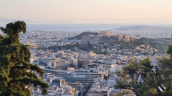 Panoramaaufnahme der Stadt Athen mit den verschiedenen Gebäuden sowie der Sehenswürdigkeit Akropolis. Im Hintergrund ist das Meer