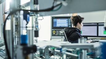 Ein junger Mechatroniker arbeitet am Computer. Im Vordergrund Elektronik und Maschinen