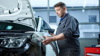 Ein Mann steht an einem Auto mit offener Kühlerhaube, dessen vorderes Heck samt Reifen abmontiert sind