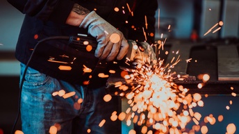 Eine Person mit dicken, grauen Arbeitshandschuhen, Jeans und dunklem Oberteil beugt sich über eine Arbeitsplatte und führt Schweißarbeiten durch. Funken sprühen ringsum.