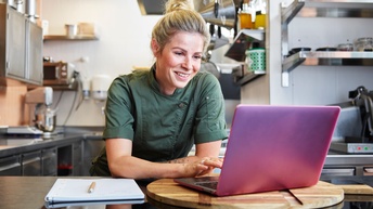 Person mit blonden geschlossenen Haaren und dunkelgrüner Arbeitsbekleidung steht in einer Gastronomieküche bei einer Küchenfront angelehnt und blickt freudig auf einen rosafarbenen Bildschirm eines Laptops, daneben liegt ein Notizbuch mit Stift