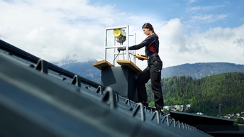 Eine Frau arbeitet an einem Kamin. Im Vordergrund das schwarze Schrägdach. Im Hintergrund eine Bergkulisse