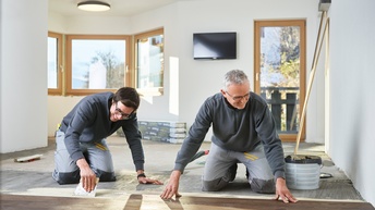 Zwei Personen in grauen Arbeitsmonturen knien am Boden und verlegen Holzbodenbretter, im Hintergrund Fenster, Balkontüre und Fernseher