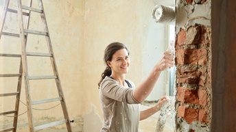 Eine junge Frau streicht mit der Rolle eine Mauer im sanierten Altbau. Im Hintergrund steht eine Leiter