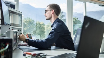 Ein junger Mann mit Messgerät sitzt im Büro an einem Computer. Im Vordergrund ein offener Laptop