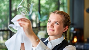 Lächelnde Person mit Zopf in grauem Gilet und weißem Hemd hält leere Weinglas mit weißem Tuch umfasst in die Luft und blickt darauf
