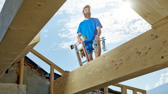 Blick von unten auf eine Person mit blauem T-Shirt, die auf Leiter steht und zwischen Holzbalken hervorlugt 