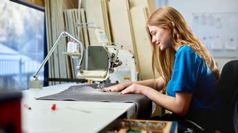 Eine junge Frau näht an einer Nähmaschine. Im Hintergrund große Holzteile