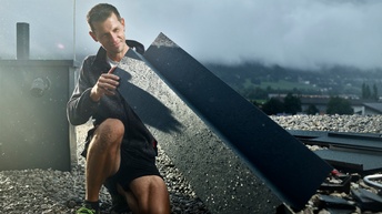 Ein Spengler auf einem Flachdach betrachtet ein Blechteil zur weiteren Verarbeitung. Im Hintergrund eine nebelige Landschaft