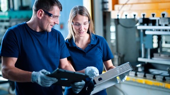 Zwei Personen mit Schutzbrillen und Handschuhen halten metallene Objekte, im Hintergrund verschwommen Industriemaschinen